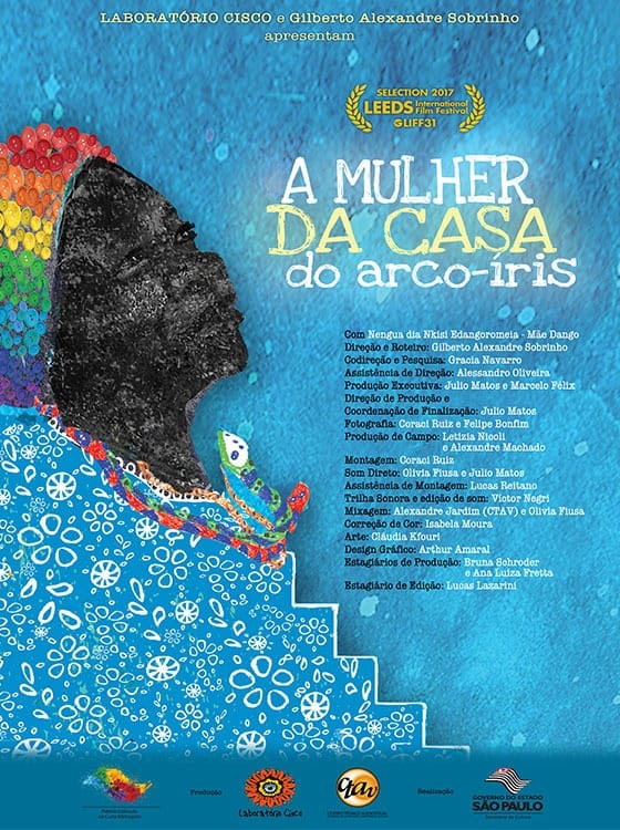 A imagem apresenta um cartaz de filme, com o fundo azul com uma mulher negra utilizando uma touca feita de botões coloridos e um vestido azul com flores abstratas brancas, o contorno do vestido traz o formato de escada. No pescoço da mulher tem dois colares em formatos de cobras, uma verde e a outra vermelha. No centro o nome da obra “A mulher da casa do arco-íris”, abaixo o nome da produção [ Com: Nengua dia Nkisi Edangoromeia - Mãe Dango; Direção e Roteiro: Gilberto Alexandre Sobrinho; Codireção e Pesquisa: Gracia Navarro; Assistência de Direção: Alessandro Oliveira; Produção Executiva: Julio Matos e Marcelo Félix; Direção de Produção e Coordenação de Finalização: Júlio Matos; Fotografia: Coraol Ruiz e Felipe Bonfim; Produção de Campo: Letizia Nicoli e Alexandre Machado; Montagem:Caraol Ruiz; Som Direto: Olivia Fiusa e Julio Matos; Assistência de Montagem: Lucas Reitano; Trilha Sonora e edição de som: Victor Negri; Mixagem: Alexandre Jardim (CTAV) e Olivia Fusa; Correção de Cor: Isabela Moura; Arte: Cláudia Kfourl; Design Gráfico: Arthur Amaral; Estagiários de Produção: Bruna Sohroder e Ana Luiza Fretta; Estagiário de Edição: Lucas Lazarini.] Na parte superior temos o texto “Laboratório Cisco e Gilberto Alexandre Sobrinho" e na parte inferior os patrocinadores do projeto da obra.