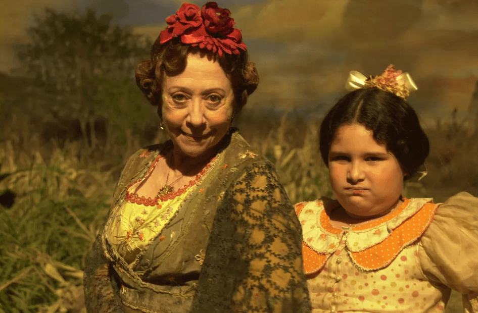 Fotografia parcial de Fernanda Montenegro e uma atriz mirim interpretando personagens. Ambas estão olhando com expressões de seriedade em direção a câmera. Fernanda utiliza flores violetas na cabeça e um penteado clássico de época, além de um vestido verde com muitos detalhes bordados e uma manta fina, também com detalhes bordados, jogadas sob seus ombros. Em seu peito há um colar de cor ouro, grande e cheio de detalhes. A menina ao lado esta com seus cabelos negros presos e um laço bege com flores rosas no topo da cabeça. Ela usa um vestido com várias estampas em tons de laranja e rosa. No fundo da imagem há árvores e grama desfocadas. Toda a fotografia traz um tom amarelado antigo, dando uma sensação de envelhecimento.